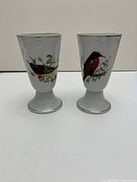 Front view of two tall handleless coffee cups featuring colorful bird motifs on white porcelain with gold trim.