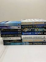 Photo shows 15 Louise Penny books arranged in two stacks on a white surface. Seven hardcover books stack on the right, eight paperback books on the left.