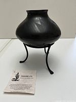 Black crafted and etched pottery vase on black metal stand shown with authenticity card