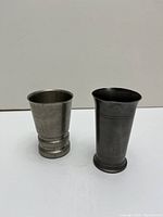 Front view of two metal cups, pewter on left, darker tin cup on right