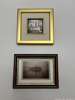 Two framed art pieces: top one is a small framed ceramic painting of a Moscow city view in gold-colored frame; bottom one is a framed print of a spire on an island in a lake in a wooden frame.