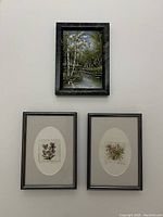 Three small framed artworks including two floral prints with gray metallic frames and one green landscape in relief in stone frame on wall