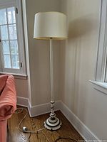 Tall painted floor lamp with white barrel shade and two-tone decorative base with stone accent detail, placed next to a window and a peach sofa on hardwood floor.