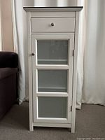 Front view of the tall narrow white cabinet showing the top drawer and lower door with frosted glass panels.
