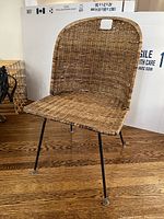 Photo of a brown wicker chair with metal legs on a wooden floor, showing the full seat and backrest made of wicker and the black metal leg structure.