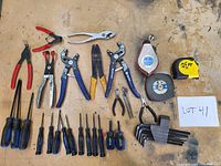 Photo showing assorted screwdrivers with blue/black handles, multiple pliers, two tape measures, and a set of black hex keys on a wooden surface