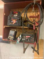 Battery booster pack with attached jumper cables, battery charger unit, and analog load tester meter on wooden shelving.