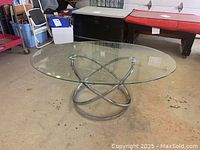 Full view of oval clear glass top table with intertwined polished chrome circular base on a concrete floor.
