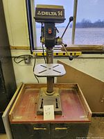 Delta 14-inch drill press mounted on a wooden cabinet with storage doors, showing the front view of the tool and cabinet.