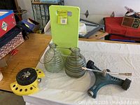 Photo showing two vintage ribbed glass garden sprayers, yellow circular sprinkler, and lime green foam kneeling pad on white tablecloth.