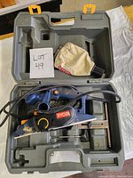 Open gray plastic carrying case with Ryobi electric hand planer inside, along with a dust collection bag and control accessories.