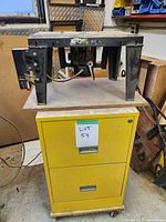 Photo showing a router table mounted on a yellow two-drawer cabinet with wheels, power switch and power cord visible underneath router table.
