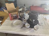 Two fondue sets displayed on a white tablecloth. One set is stainless steel with an electric heater and forks, the other is black cast iron with a stand, pot, forks, and white ceramic bowls. Original box for stainless steel set included.