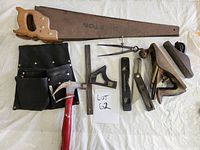 Image of two wooden hand saws, black tool belt, claw hammer, square ruler, marking and bevel gauges, wooden and metal hand planes and dividers, laid out on fabric.