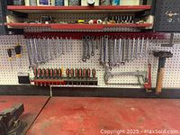 Workshop pegboard with assortment of wrenches of different sizes, nut drivers, hacksaw, and hammer hung up above a red workbench. Bench vice is visible but not included in the lot.