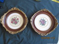 Both framed petitpoint floral needlework pictures side by side on a dark surface, showing the overall round ornate frames and needlework designs.