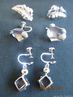 Three pairs of sterling silver clip-on earrings arranged on black fabric: leaf-shaped, flower-shaped, and black geometrical dangle styles.