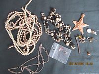 Photo showing multiple costume jewelry items including pearl-style necklaces, beaded black and pearl necklace, chain necklace, wooden star ornament, assorted small jewelry parts and a plastic bag with small colored gemstones.