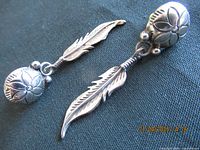 Front view of two feather earrings laid flat on blue fabric showing floral top and feather pendant detail.