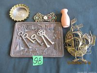 Photo showing entire lot with all items: brass ashtray, metal brooch, wooden vase, decorative brass ship, Colorado souvenir tray with skeleton keys on it, and small label with number 98.