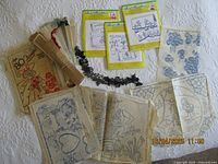 Photo showing a collection of vintage needlework transfer sheets, packages of Aunt Martha's embroidery transfers, a cardboard 80 designs booklet, and a black lace trim on a white surface.