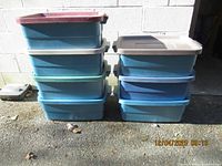Stack of eight Rubbermaid plastic storage totes in various colors with lids shown outdoors against a cinder block wall under daylight.