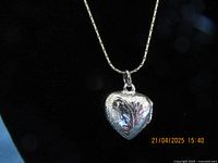 Heart-shaped silver locket on a black background showing leaf engraving detail.