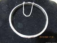 Full view of silver bangle bracelet laid on black velvet showing circular shape and safety chain attachment.