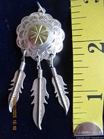 Front view of silver pendant with scalloped edges, central circular raised design, and three hanging silver feather charms. Measuring tape alongside for scale shows about 2 inches in length including feathers.