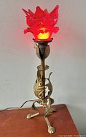 Front view of the lamp illuminated showing red floral glass shade and metal leaf base with claw feet.