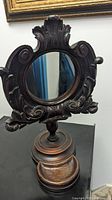 Front view showing ornate carved wood frame with circular mirror and base stand.
