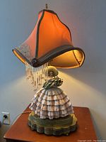 Front angled view of the lamp illuminated, showing porcelain figure and silk shade with bead fringe.