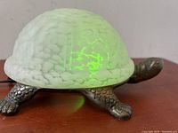 Side view of the vintage turtle lamp showing the white and green stained glass shell glowing green and metal legs on wooden surface.