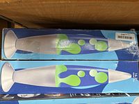 Two boxed blue lava lamps with green wax visible on the box artwork, boxes show rack wear and creases but lamps are new in box.