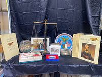 Wide view of the lot showing all items including books, plates, scale, ornament, and framed photo print arranged on black fabric.