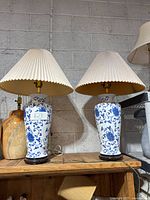 Pair of blue and white ceramic table lamps with wooden bases and beige pleated shades on a wooden surface with concrete wall background.