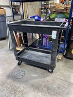 Black heavy-duty plastic Rubbermaid rolling cart with two shelves and four caster wheels, shown from one side.