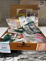 Wooden box labeled 'Elegantes' containing assorted pins in packaging and loose.