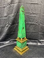 Obelisk standing on a black cloth background showing vivid green faux malachite enamel with brass base and tip detailing.