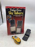 Boxed Fisher-Price Sky Talkers walkie talkies along with a yellow Motorola walkie talkie and a black Motorola flip phone on a white surface.