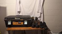 Bostitch toolbox, Carolina boots, and Vans sneakers displayed on wooden table against wall.