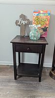 Full view of dark brown wooden side table holding the cross, glass vase, and cereal box showing size and condition