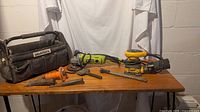 Wide shot of all items on a wooden table: tool bag, Ryobi belt sander, DeWalt orbital sander, chisels.