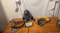 All three power tools on a wooden surface: Ridgid orbital sander, Ryobi router with dust bag, and DeWalt jigsaw.