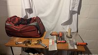 Photo showing the red wheeled suitcase/backpack and six drywall tools including putty knives and taping knives on a wooden table against white cloth and concrete wall background.