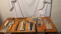 A wooden table displaying a variety of drywall tools including hammers, trowels, knives, a roller, and scraping tools, arranged in two rows.