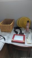 Full view of all items on table including basket, yellow hangers, soda bottle, iPhone case, black collar with fan, wallet, cross, Etch A Sketch, and black curved item.