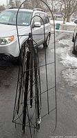 Black metal garden trellis leaning against a car in a driveway on an icy day, showcasing grid pattern and pointed top end.