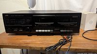 Front view of JVC TD-W118 double cassette deck showing both cassette compartments, control buttons, power switch, input level knob, and Dolby B noise reduction label. Unit is black and appears intact with cables attached.