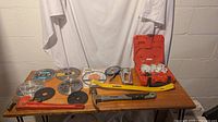 Tools and blades laid out on a wooden surface showing various circular saw blades, hammer, staple gun, yellow pry bar and the open Milwaukee carrying case.